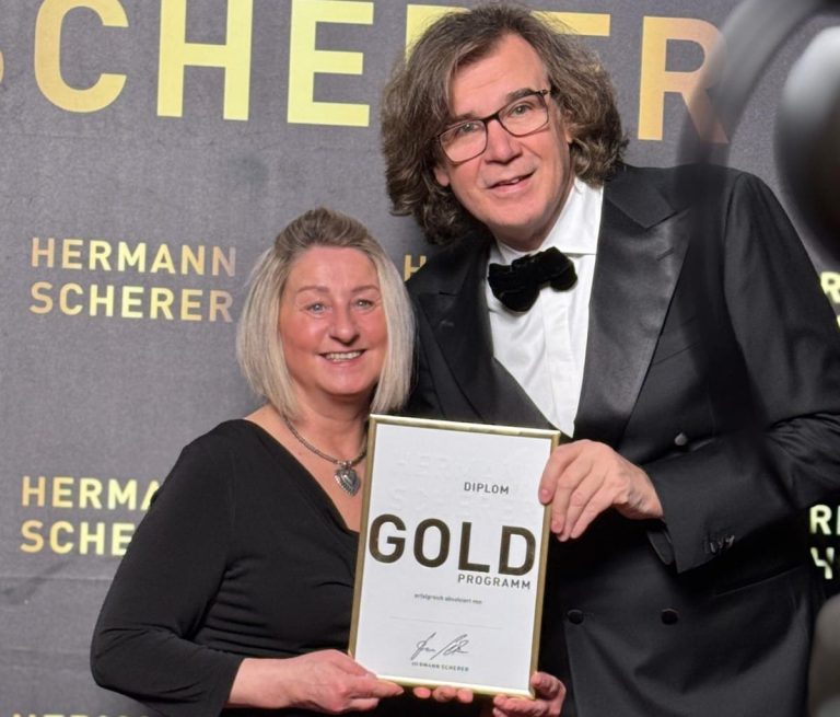 Ingrid Angelika Müller mit Hermann Scherer Zwei Personen in formeller Kleidung halten eine Auszeichnung vor einem dekorativen Hintergrund.