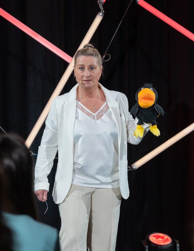 Ingrid Angelika Müller beim Speaker Slam Eine Frau in einem weißen Blazer hält einen Puppenvogel und spricht zu einer Gruppe.
