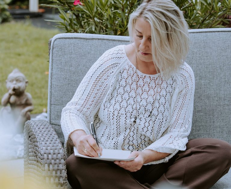 Ingrid Angelika Müller Frau in weißer Strickbluse sitzt und schreibt in einem Notizbuch, umgeben von Pflanzen.