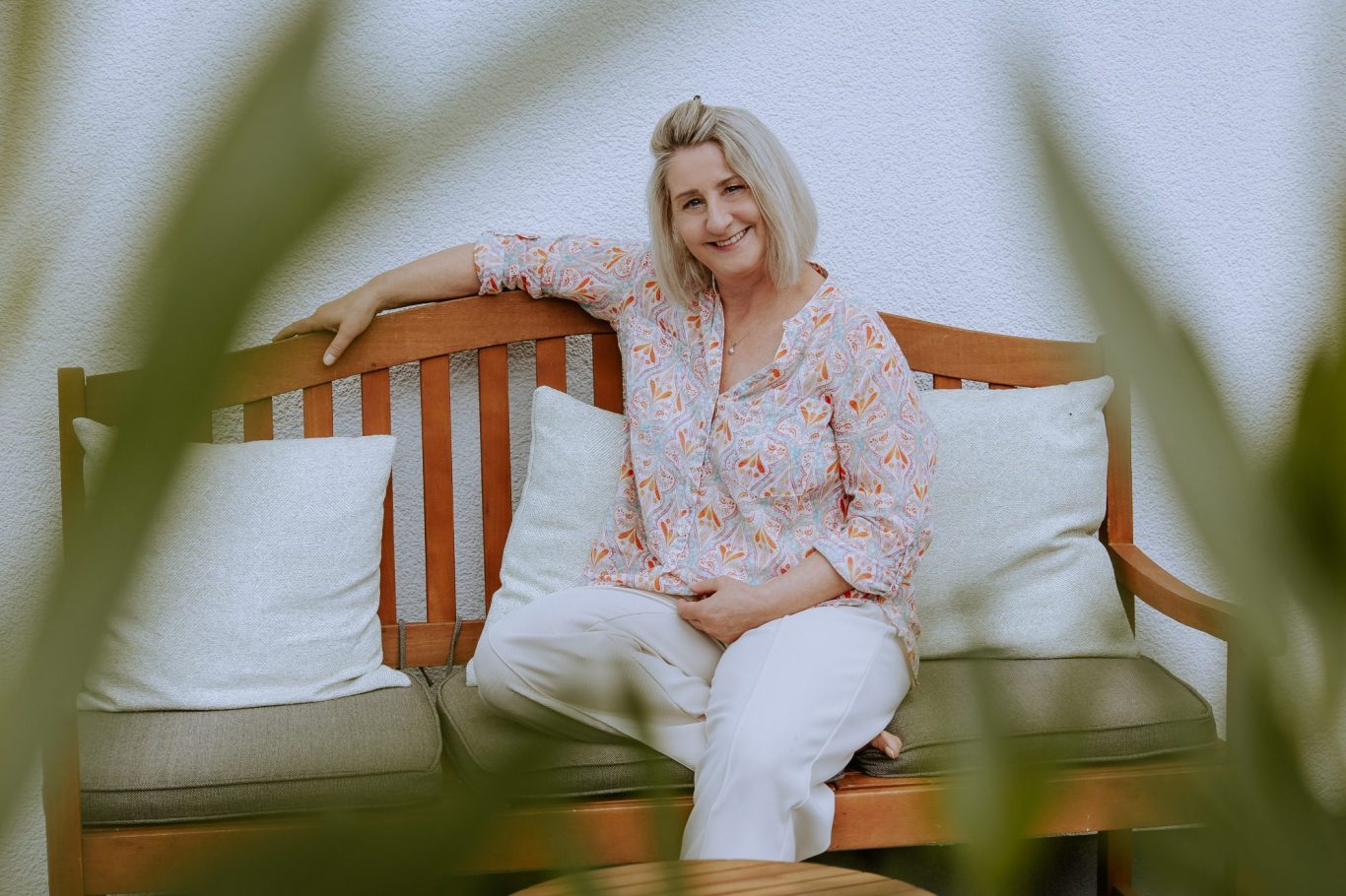 Ingrid Angelika Müller Frau mit blonden Haaren sitzt entspannt auf einer Holzbank zwischen Kissen.